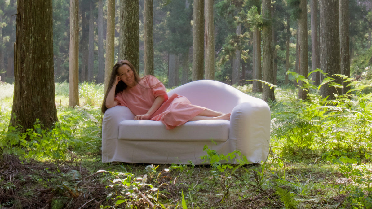 A woman in a pink robe reclines on a white armchair outdoors in a dense forest with tall trees and green undergrowth.